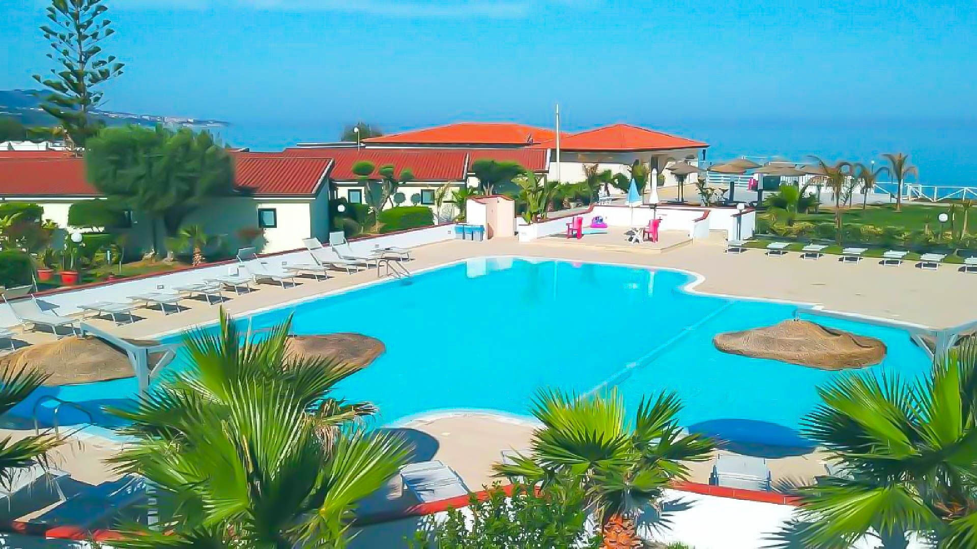 Piscina azzurra con lettini bianchi e palme tra casette dai tetti rossi, mare sullo sfondo, Borgo Marino Albatros