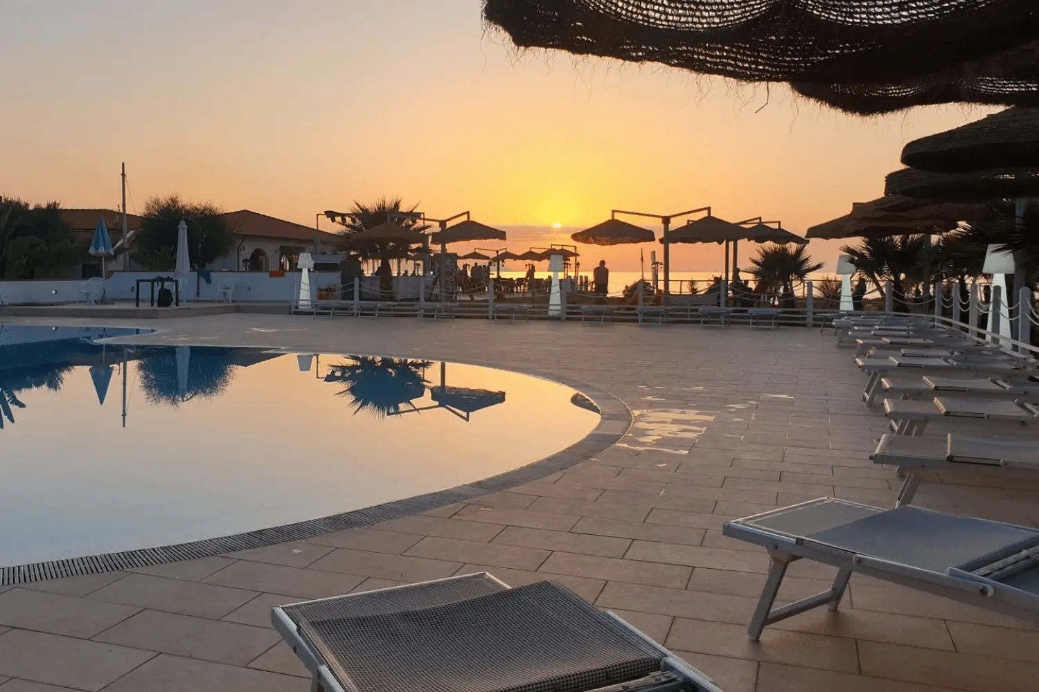 Piscina al tramonto con palme riflesse, lettini grigi e ombrelloni di paglia sul mare del Borgo Marino Albatros
