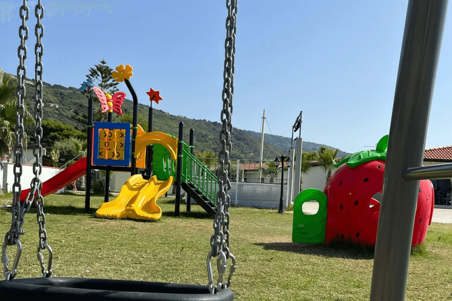 Area giochi del Borgo Marino Albatros su prato con altalena in primo piano, scivolo giallo, casetta a fragola e colline verdi